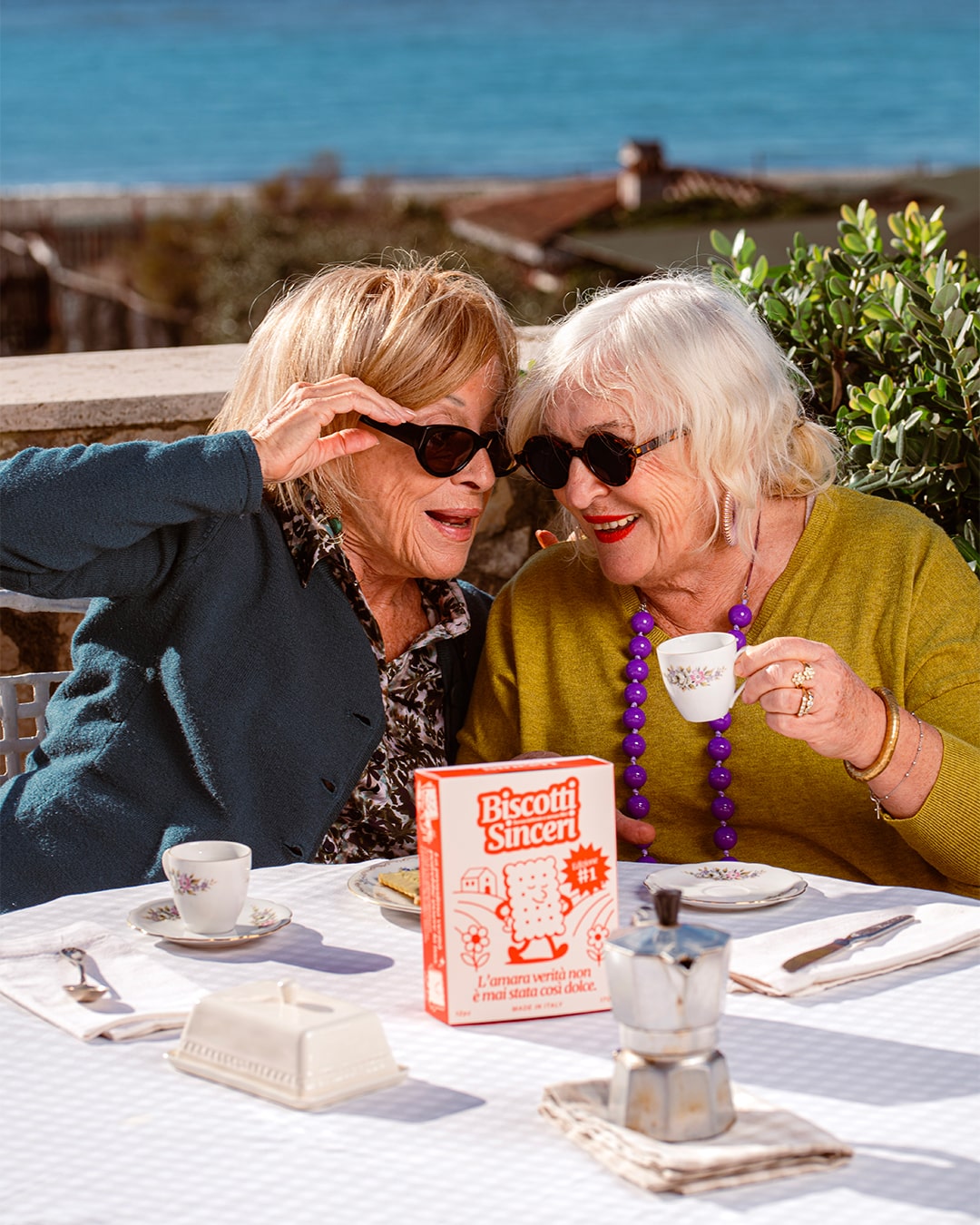 Due signore con gli occhiali da sole ridono a un tavolino sul mare, davanti a loro le tazzine del caffè e una confezione di Biscotti Sinceri